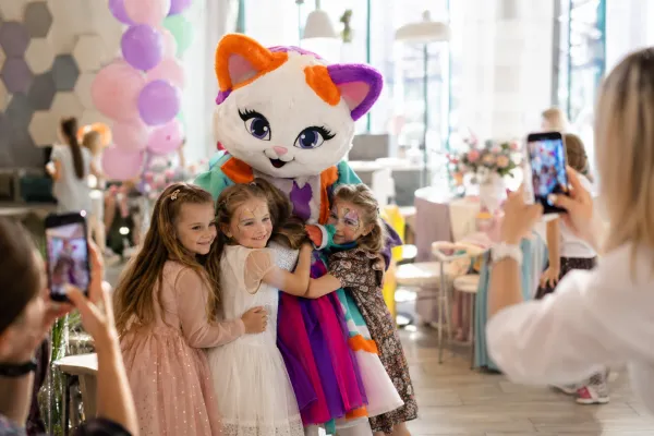 аниматор в ярком костюме кошечки фотографируется с детьми на празднике, родители снимают на телефоны